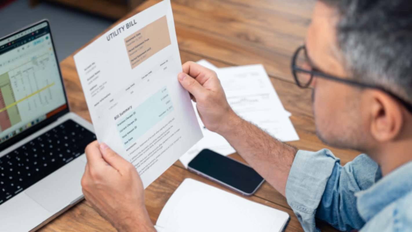 A person examines a utility bill while sitting at a desk with a laptop and a smartphone.