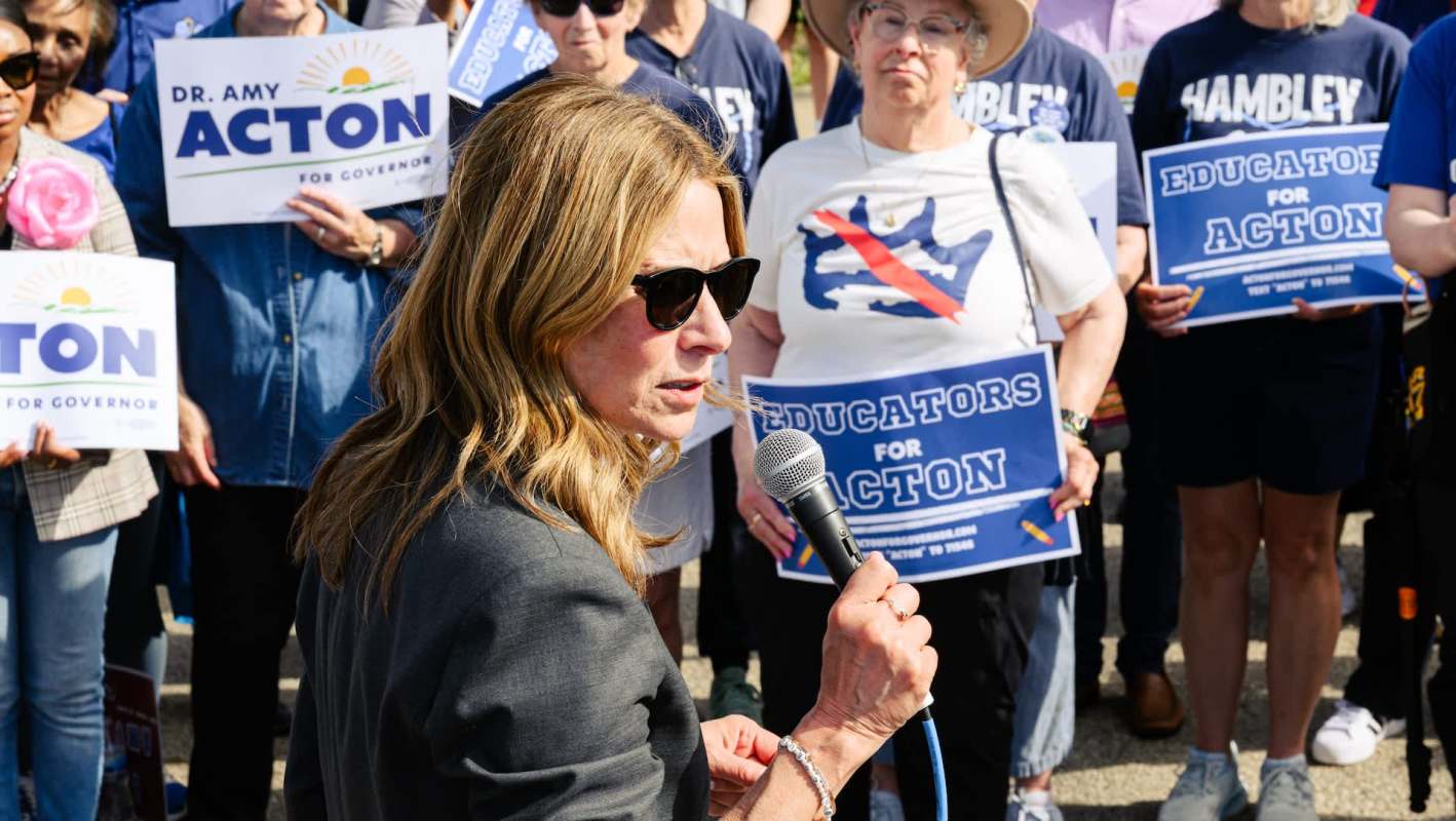Ohio Democratic gubernatorial candidate Amy Acton speaks into a microphone with supporters holding political signs.