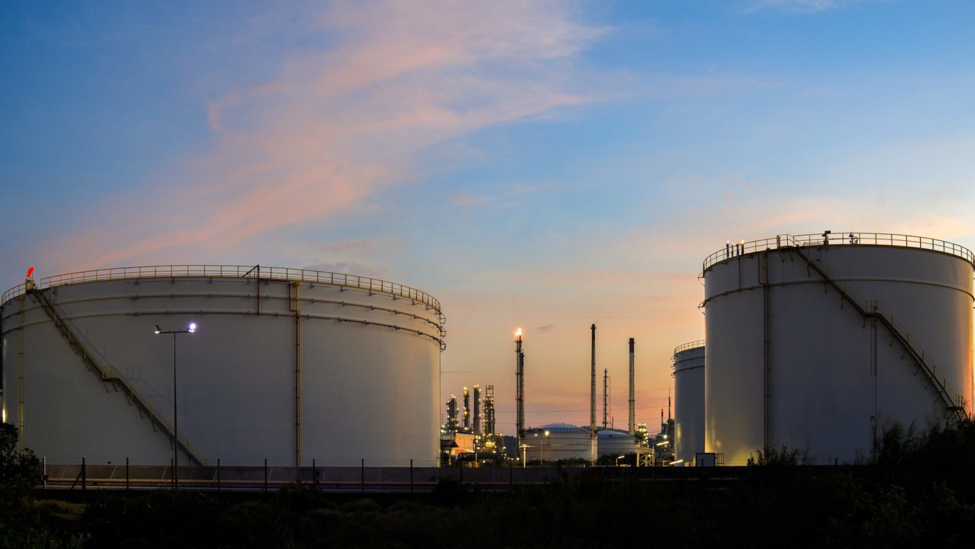 Two large industrial storage tanks are visible, with a refinery and smokestacks in the background at sunset.