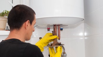 A person in yellow gloves removes a corroded part from a water heater.