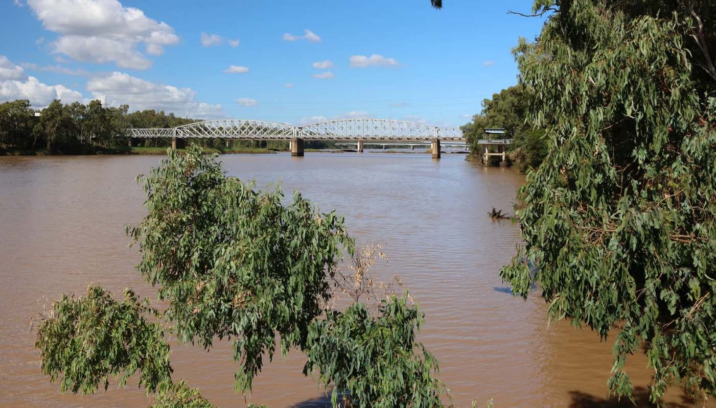 A river with brown water flows beneath a bridge, surrounded by trees and blue skies with clouds.