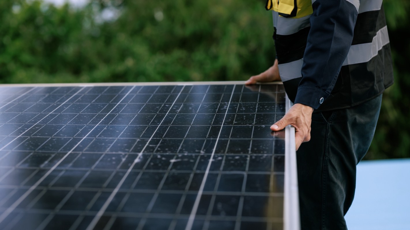 A person holds a solar panel outdoors, with greenery in the background.