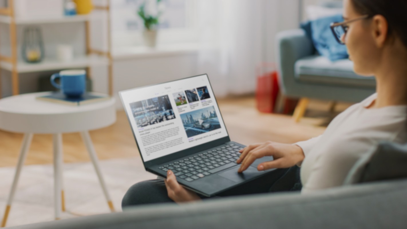 A person using a laptop while sitting on a couch in a bright living room.