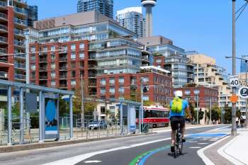 A cyclist rides on a bike lane surrounded by modern buildings and a streetcar on a sunny day.