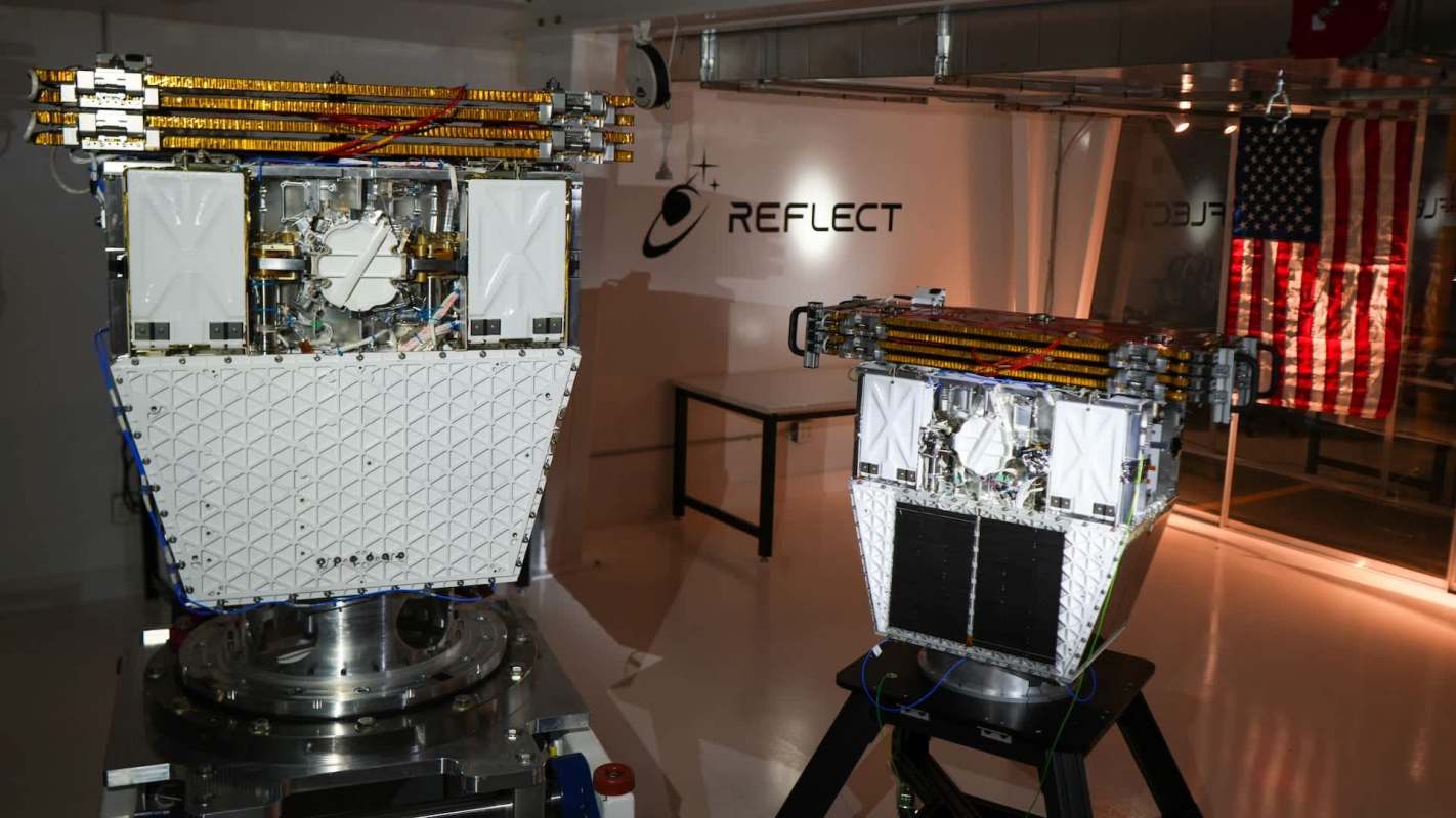 Two satellite prototypes displayed in a lab, with an American flag visible in the background.