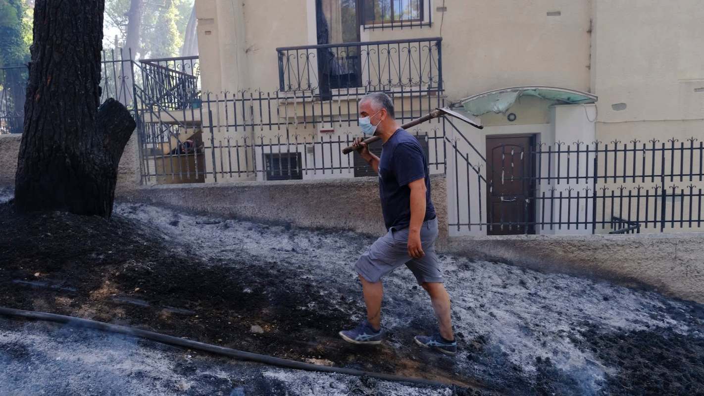 A man walks through an area of burned ground near a house, holding a tool over his shoulder.