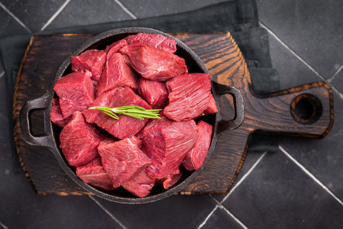 Diced raw ostrich meat sits in an iron skillet on a wooden plank topped with a sprig of greenery