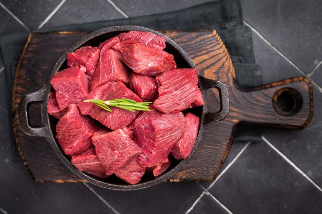 Diced raw ostrich meat sits in an iron skillet on a wooden plank topped with a sprig of greenery