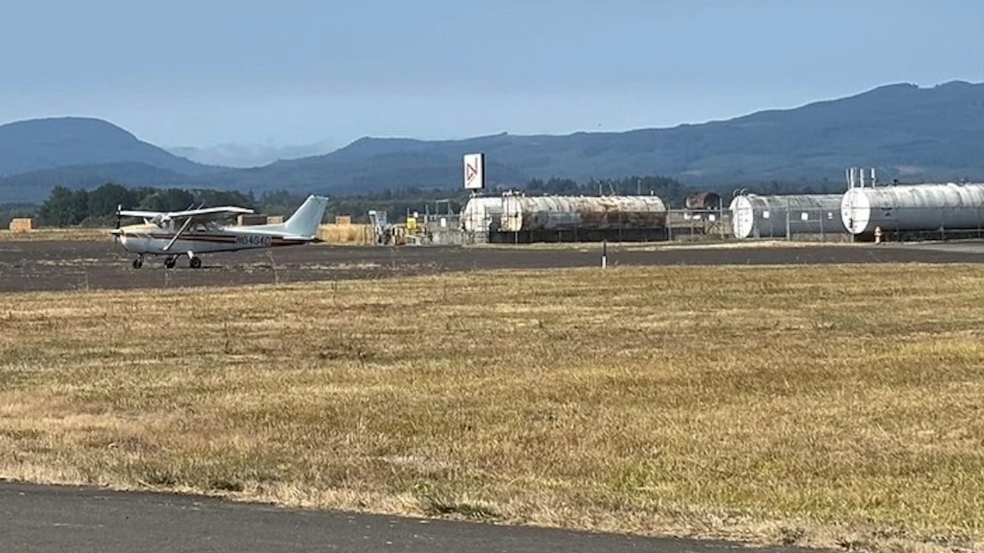 A small airplane is on a runway near storage tanks and mountains in the background.