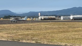 A small airplane is on a runway near storage tanks and mountains in the background.