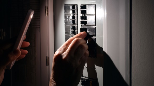 A hand adjusts a circuit breaker while holding a phone that emits light.