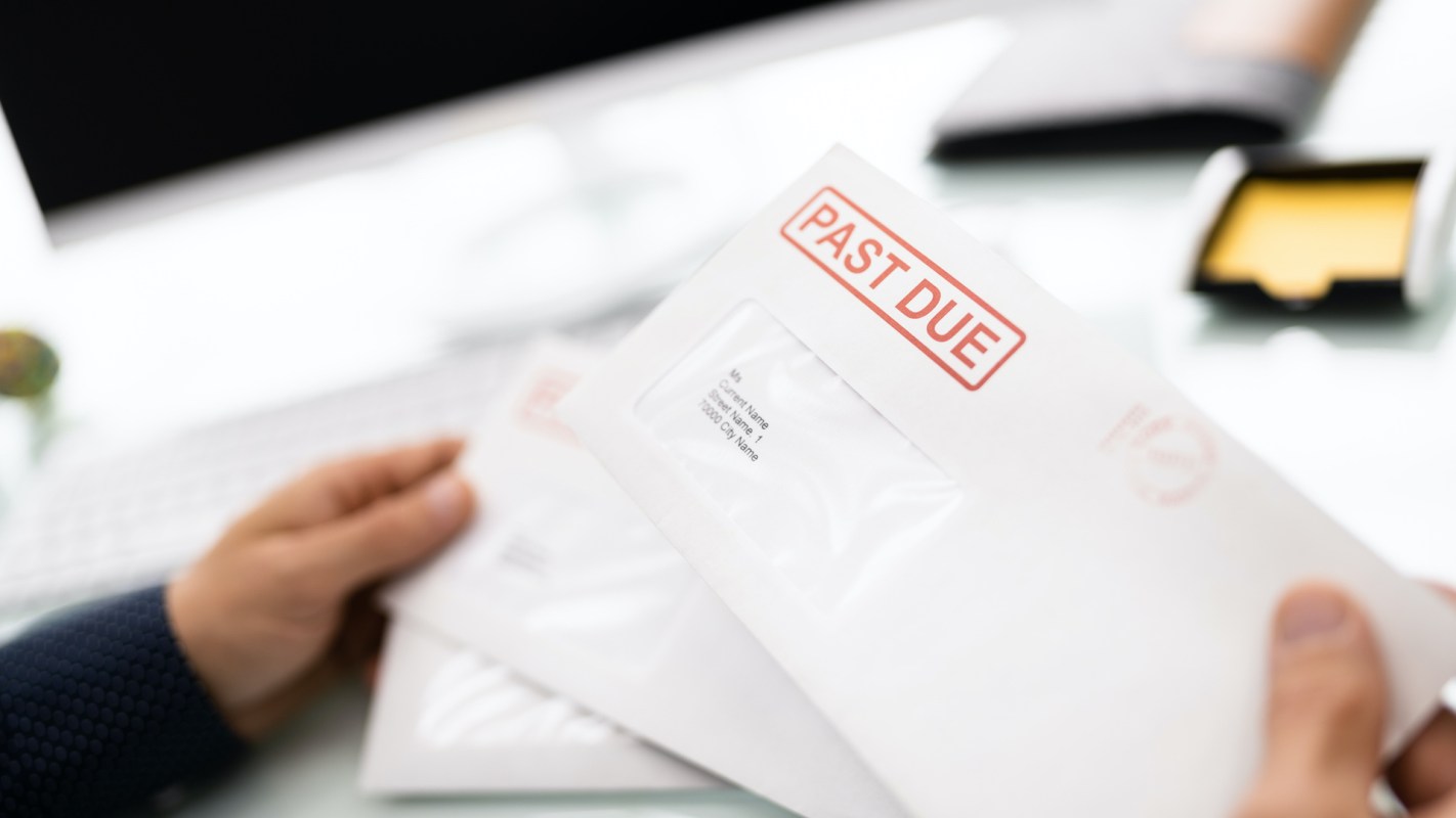 A person holding envelopes labeled "PAST DUE" on a desk with a computer in the background.