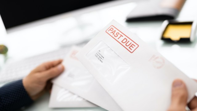 A person holding envelopes labeled "PAST DUE" on a desk with a computer in the background.
