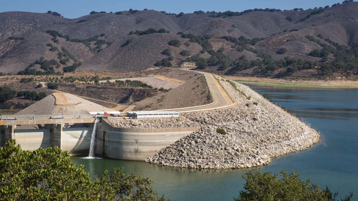 A dam structure with a spillway, surrounded by hills and a reservoir.