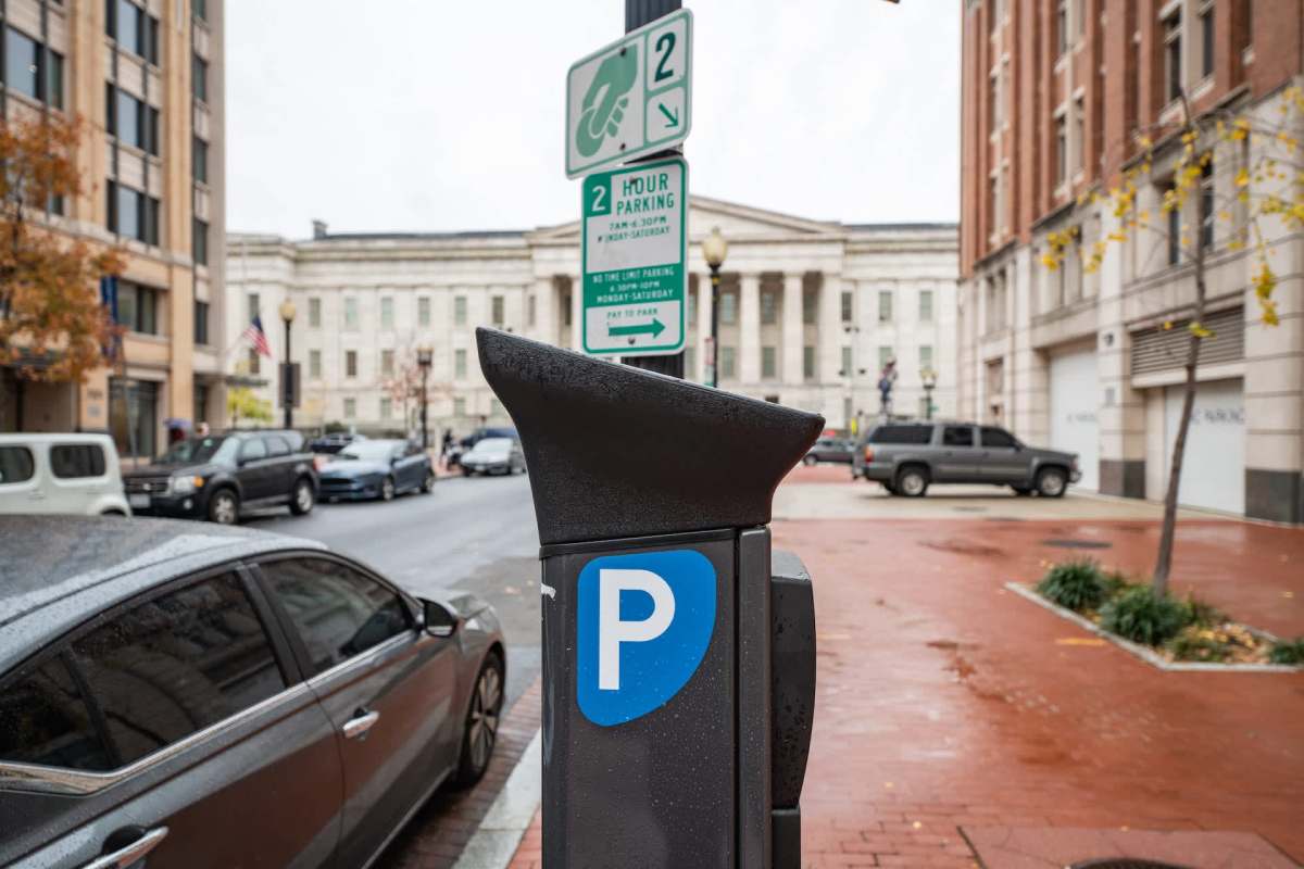 A parking meter with a "P" symbol on a city street with vehicles and buildings in the background.