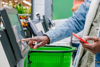 A person uses a self-checkout kiosk while holding a smartphone and a shopping basket.