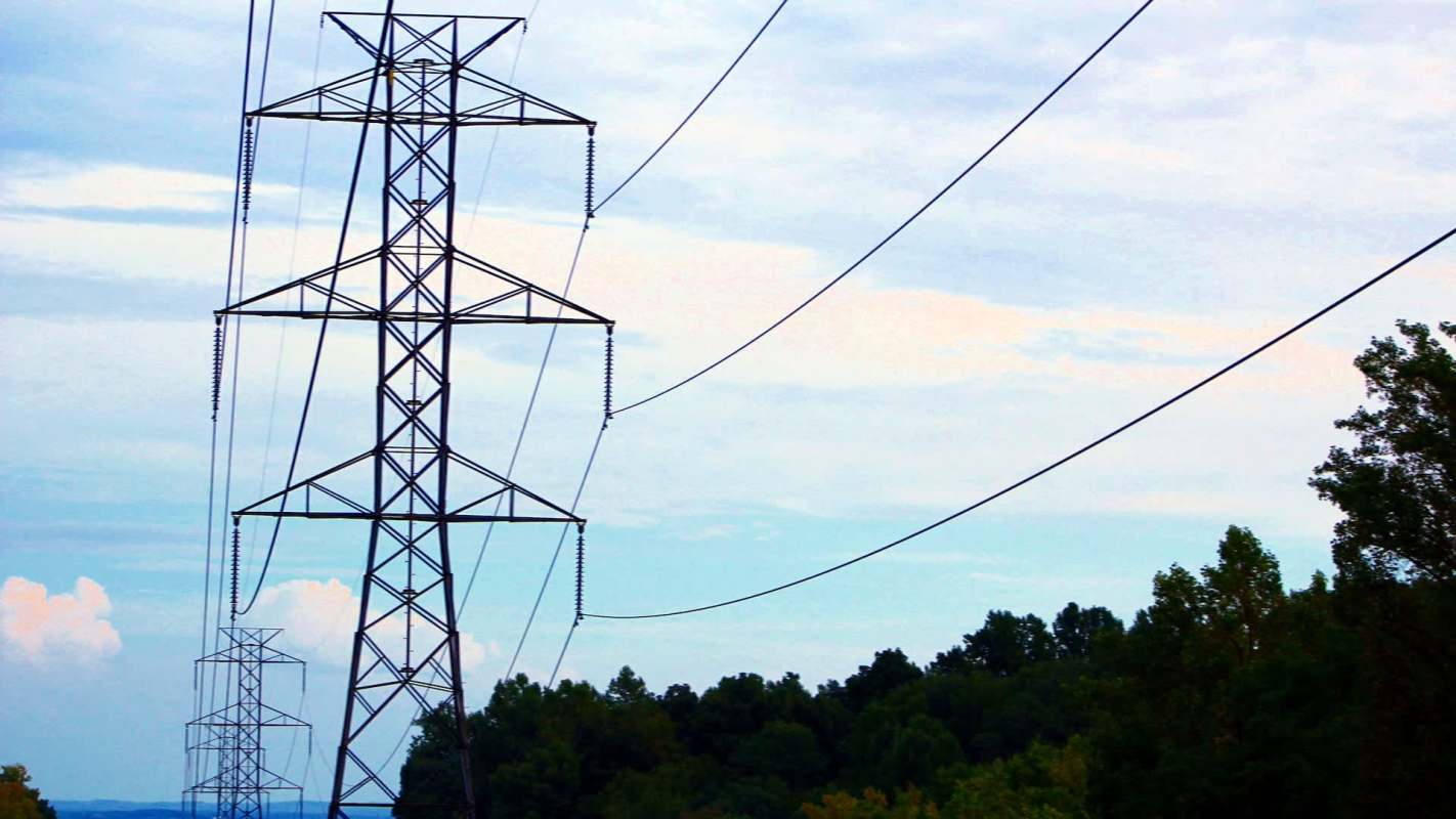 Electric transmission towers with power lines against a cloudy sky and wooded area.