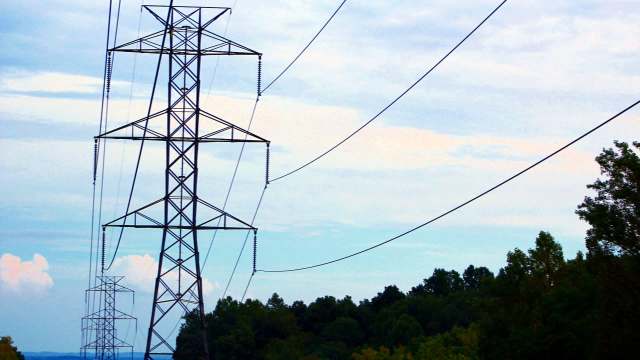 Electric transmission towers with power lines against a cloudy sky and wooded area.
