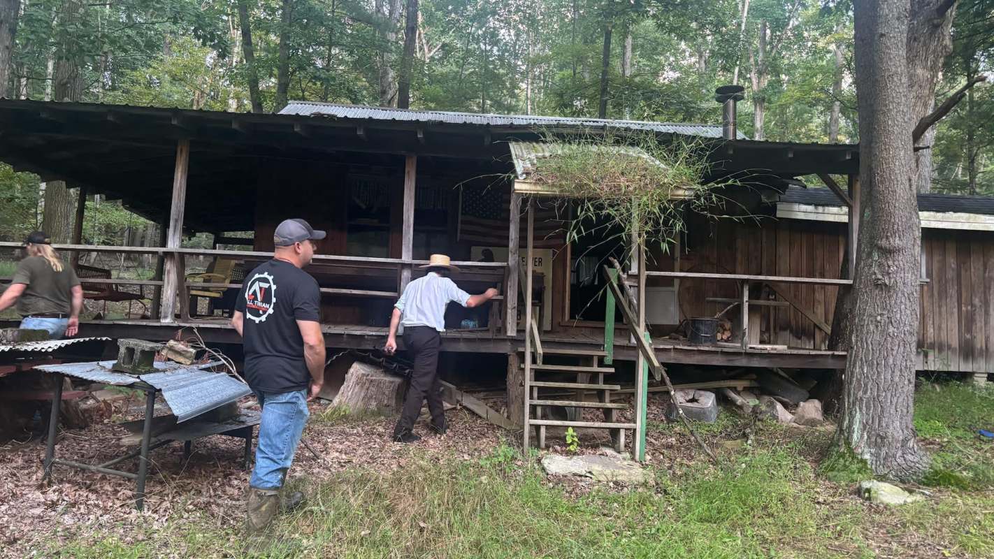 Two men approach an old wooden cabin in a wooded area, with one man ascending the steps.