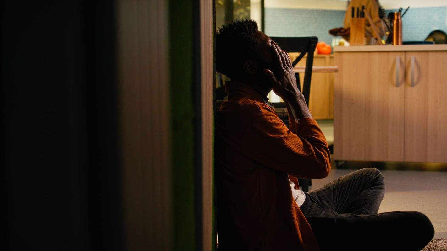 A man sitting on the ground in a hallway, with his hands covering his face.
