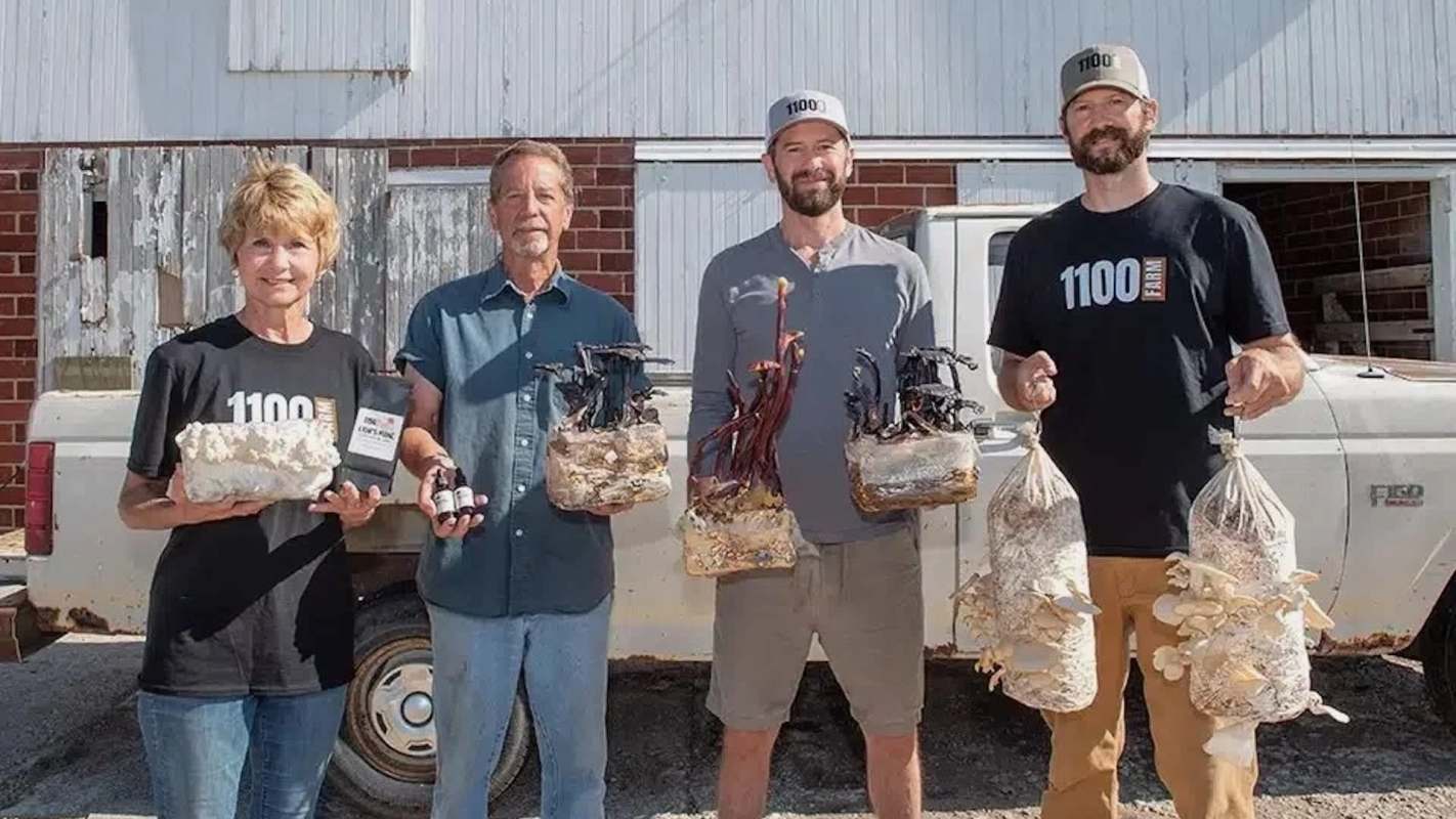 Four individuals hold various types of mushrooms and mushroom products in front of a vintage truck and barn.