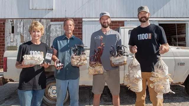 Four individuals hold various types of mushrooms and mushroom products in front of a vintage truck and barn.