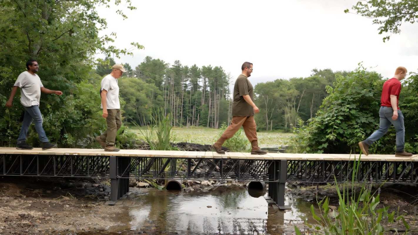Four people walk across a wooden bridge supported by a trellis over a stream surrounded by greenery.