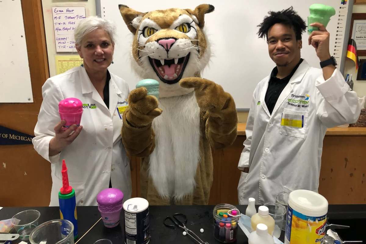 A woman and a man in lab coats pose with a mascot in a classroom, holding colorful plastic props.