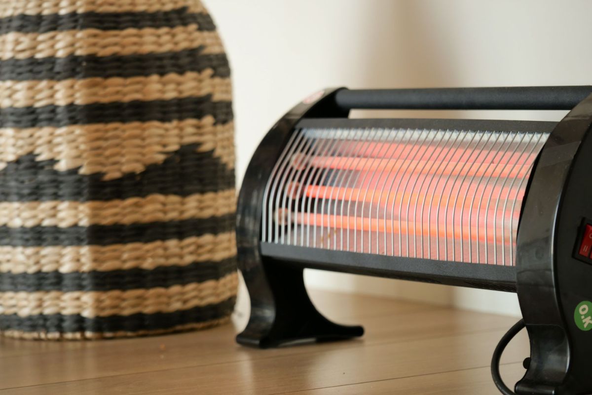 A plug in heater with a red heating element sits on a floor next to a basket
