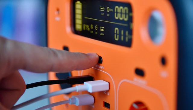 A hand presses the power button on a portable charging device with various output ports.