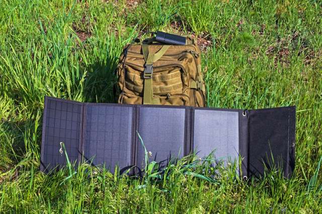 A solar panel is displayed on grass next to a military-style backpack.