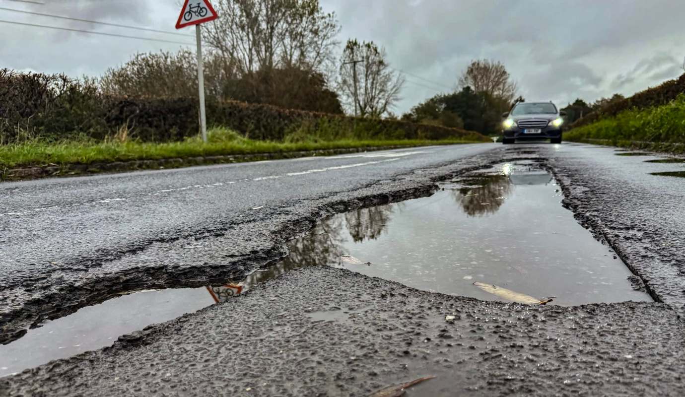 A car approaches a road with potholes and puddles on a cloudy day.