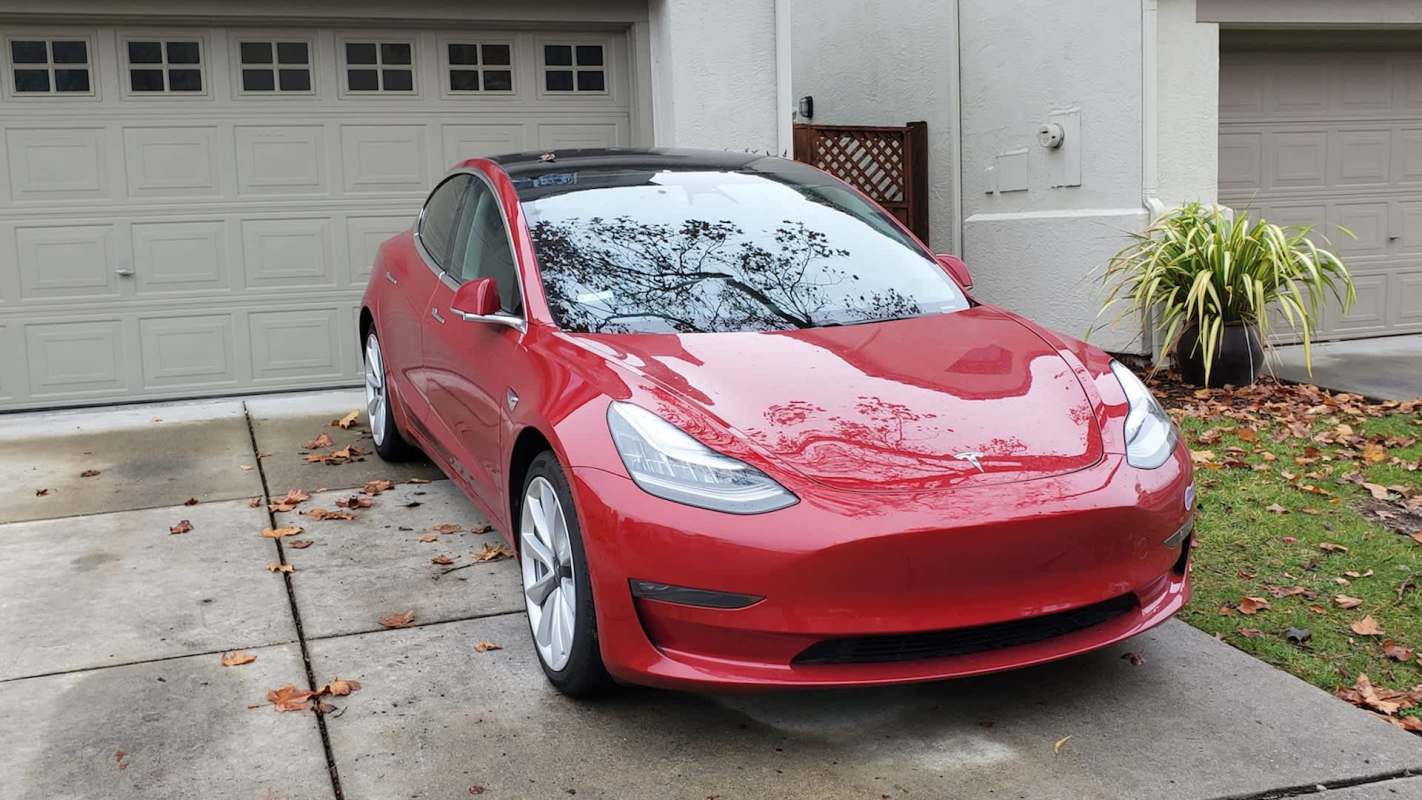 A red Tesla car parked in front of a house with a garage and scattered leaves on the driveway.