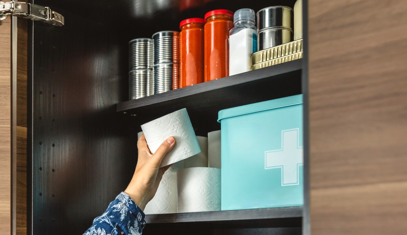 A hand reaches for a roll of paper towels on a shelf with various containers and canned goods.