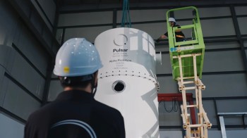 A worker on a lift installs equipment labeled "Pulsar Kinetic Energy Storage System" in a facility.