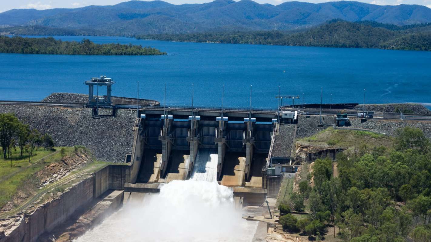 A dam releasing water into a reservoir surrounded by mountains and trees.