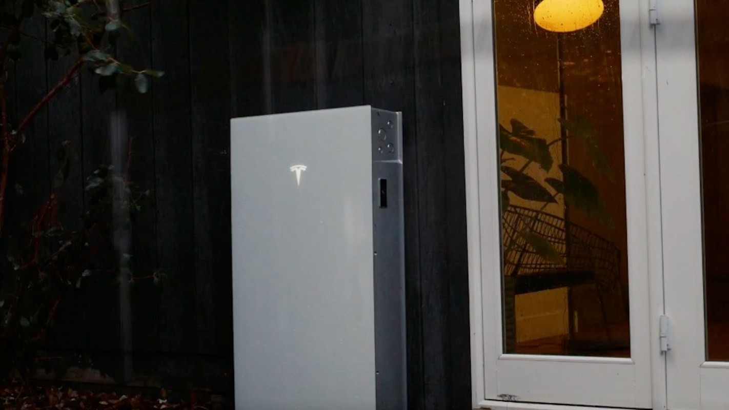 A Tesla Powerwall mounted outside a home near a window with indoor plants visible.