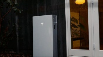 A Tesla Powerwall mounted outside a home near a window with indoor plants visible.