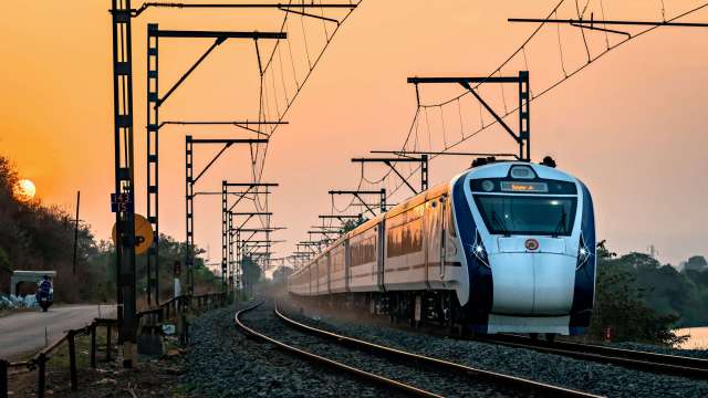 A train travels along a railway track at sunset, with power lines and roadside vegetation nearby.