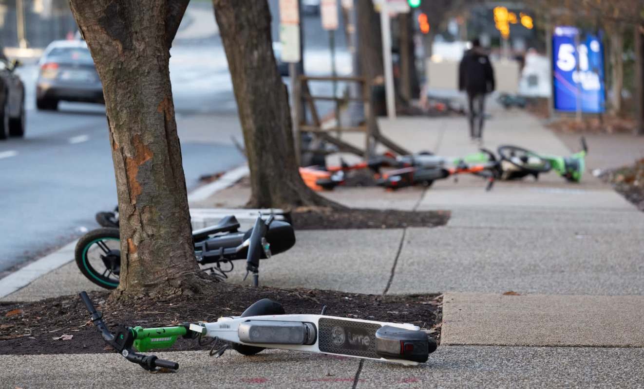 Several discarded electric scooters and electric bicycles are lying on a sidewalk near trees.