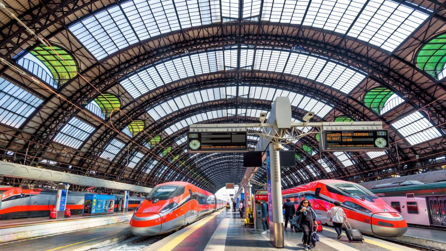 A train station with multiple trains, large arched glass roof, and digital displays showing train information.
