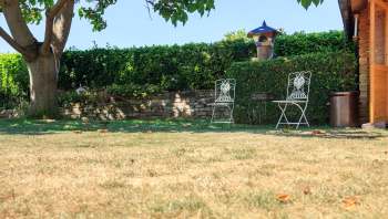 Two white metal chairs are placed on a dry lawn next to a stone wall and greenery, with a wooden structure nearby.