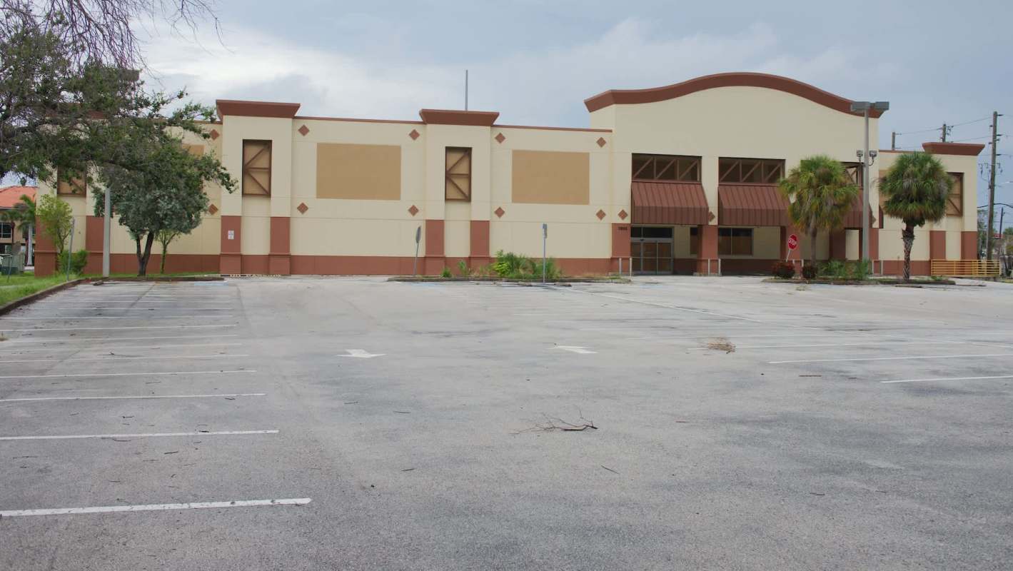 An empty building with a large parking lot and a few trees nearby.