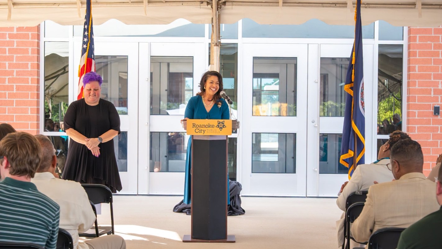 A woman speaks at a podium with the Roanoke City Public Schools sign, while another woman stands nearby.
