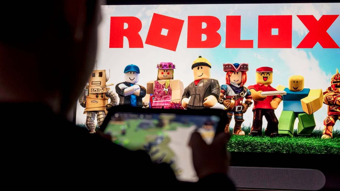 A person holds a device in front of a screen displaying characters from the game Roblox.
