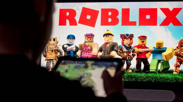 A person holds a device in front of a screen displaying characters from the game Roblox.