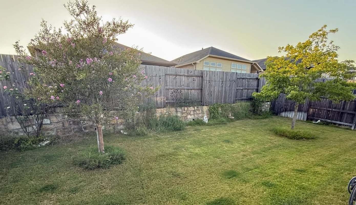 A fenced backyard with two trees, grass, and a stone wall in the background.