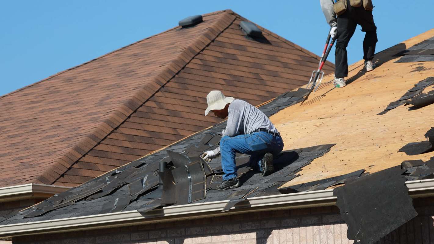 Two workers are replacing shingles on a sloped roof under a clear blue sky.