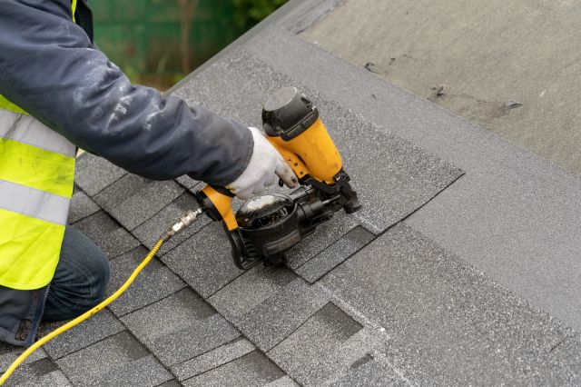 A worker using a nail gun to attach shingles to a roof.