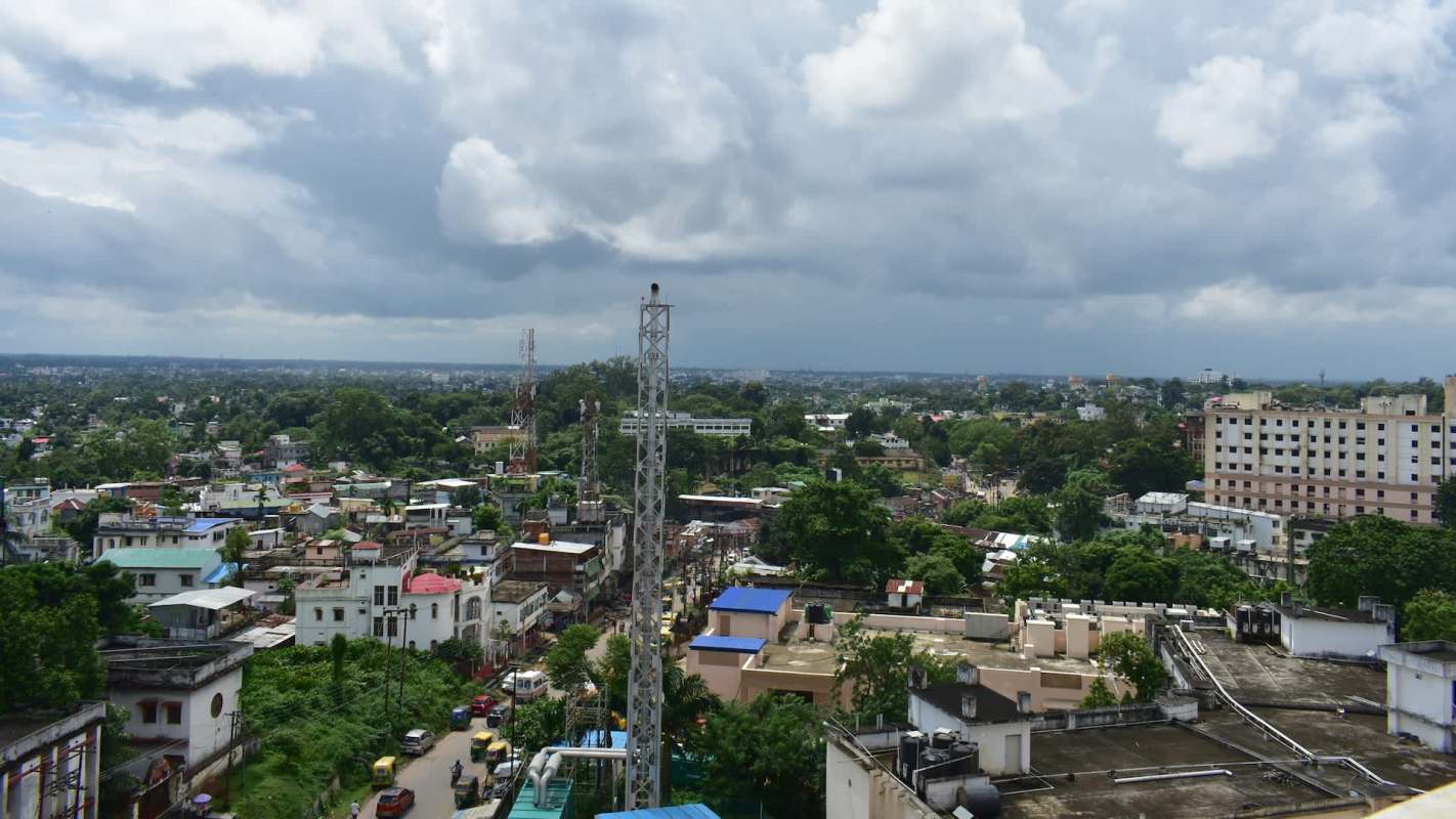 A panoramic view of a cityscape featuring buildings, trees, and telecommunications towers under a cloudy sky.